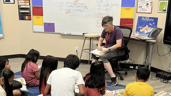 A teacher is seated in a chair reading to a group of young students who are sitting on a blue circular-patterned rug in a classroom.
