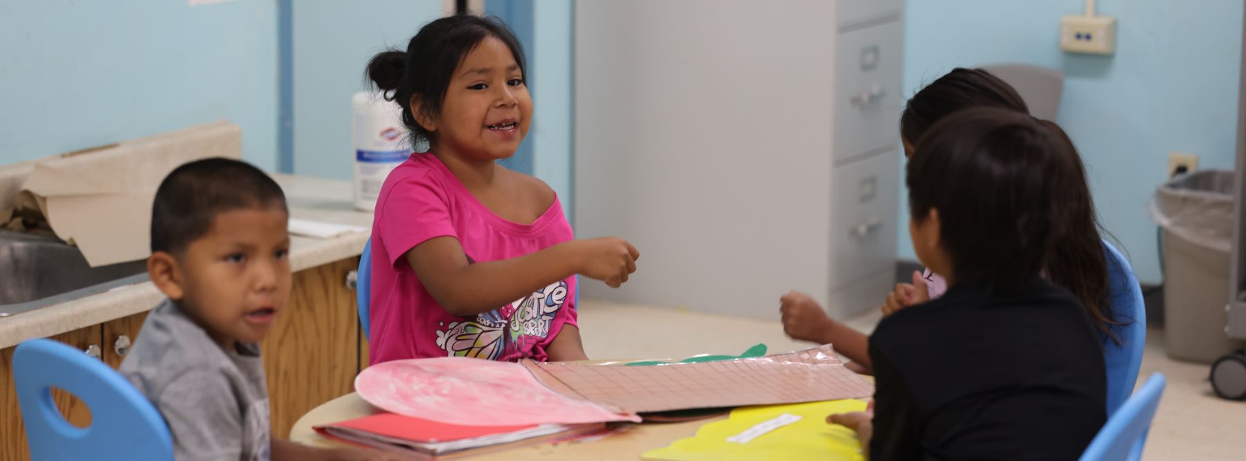 Four students sitting together at a round table with papers around and talking.