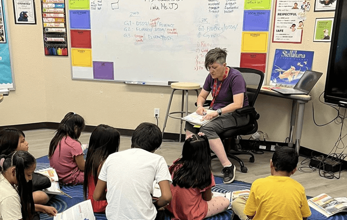 A teacher is seated in a chair reading to a group of young students who are sitting on a blue circular-patterned rug in a classroom.