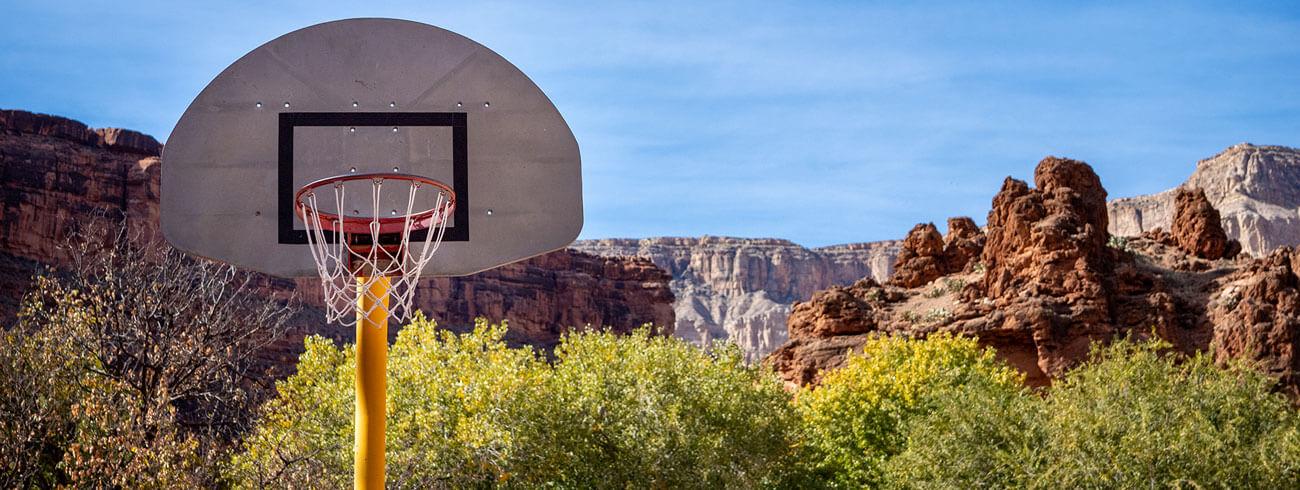 Basketball hoop in the foreground with red rock cliffs and green desert vegetation in the background under a blue sky. 