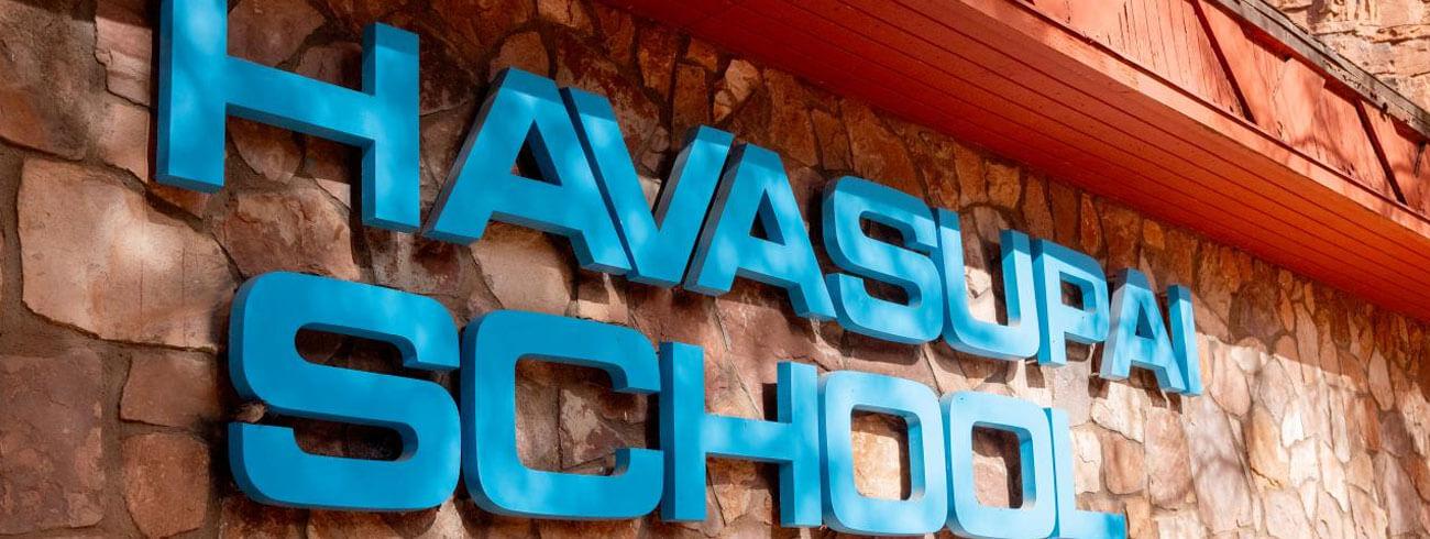 Large blue Havasupai School sign mounted on a rustic stone wall beneath a red wooden roof.